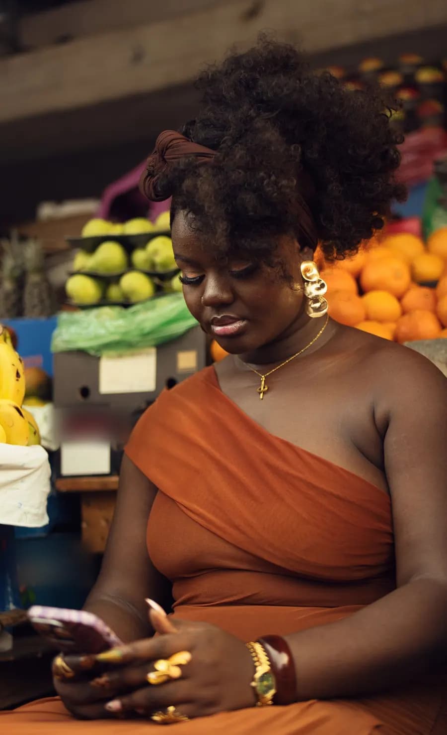 A woman in an orange dress looking at her phone at a fruit stall in a market